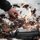 Optimus Polaris black camping stove with visible flame, used outdoors on snow and dry leaves with fuel bottle nearby