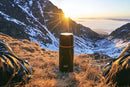 Black ESBIT stainless vacuum flask 750ml with two mugs shown outdoors in mountain sunrise setting for heat retention use.
