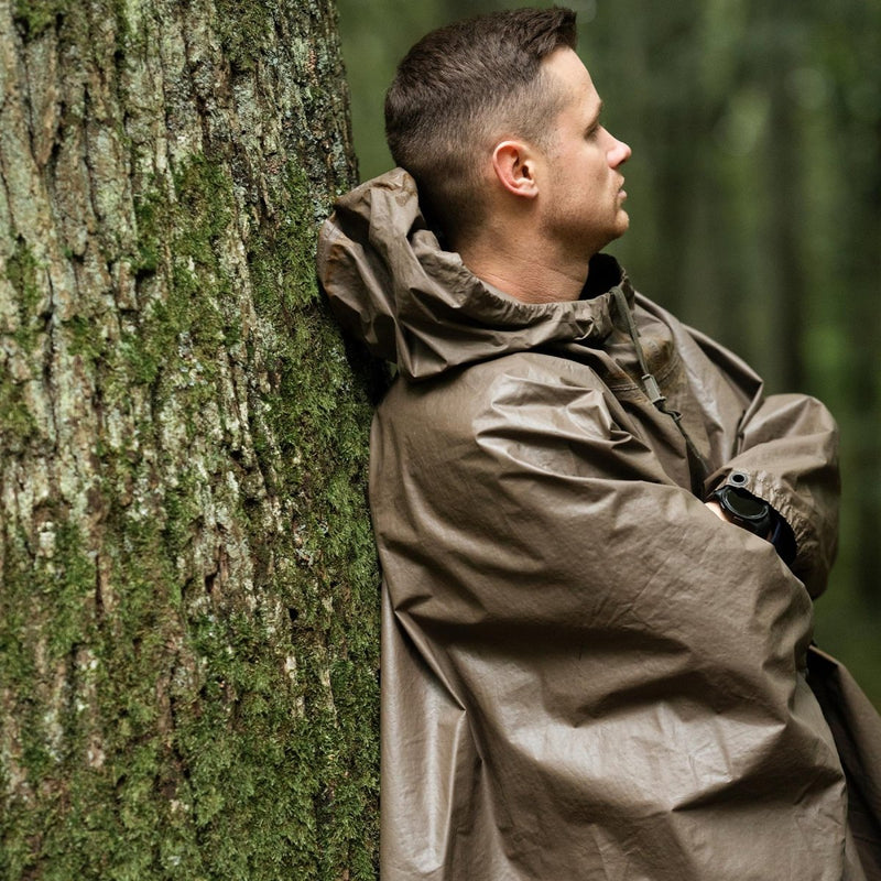 Man leaning against tree wearing a waterproof olive German army rain poncho with hood in a forest setting
