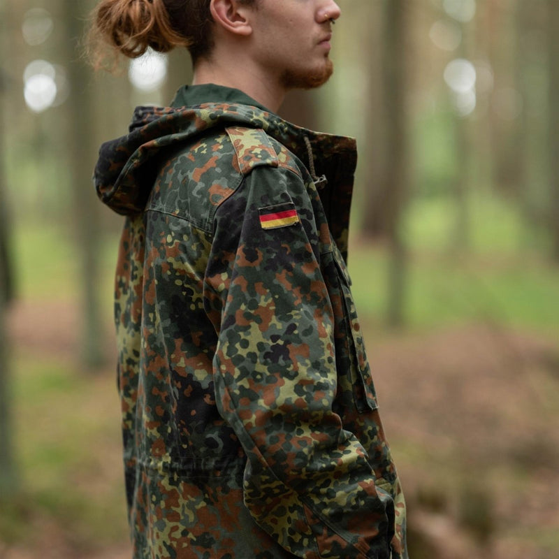 Side view of person wearing Original German army flecktarn camo hooded field jacket with German flag patch on arm outdoors